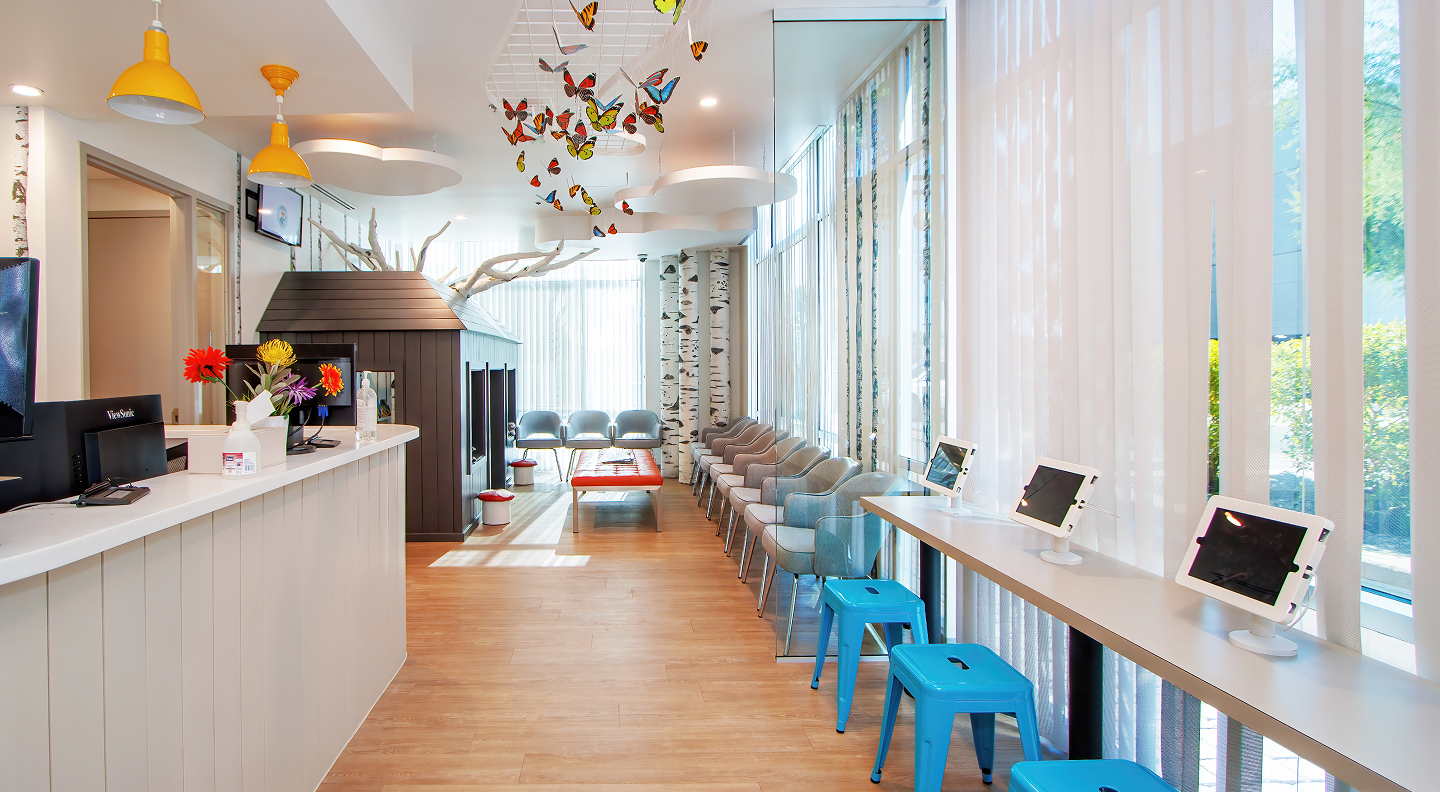Tatum Pediatric Dentistry | A modern and vibrant waiting area with a reception desk, seating, and a digital play area featuring tablets on a counter with blue stools.