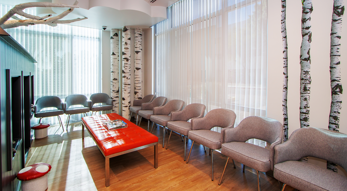 Tatum Pediatric Dentistry | A cozy waiting area with gray chairs, a red bench, and decorative birch tree wallpaper, illuminated by natural light through large windows with blinds.