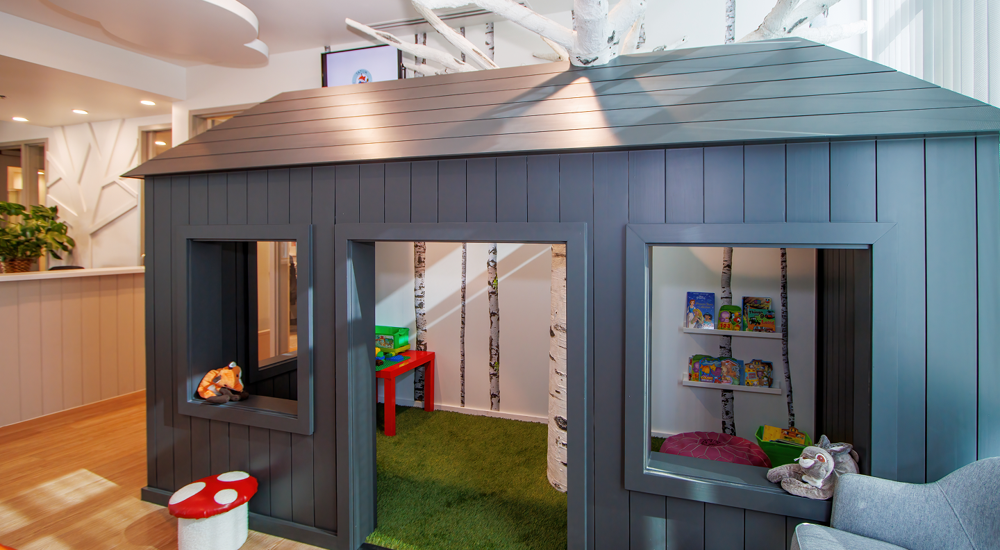Tatum Pediatric Dentistry | A children's playhouse structure in a waiting area, with a dark wooden exterior, open windows, and a green-carpeted interior stocked with books and toys.