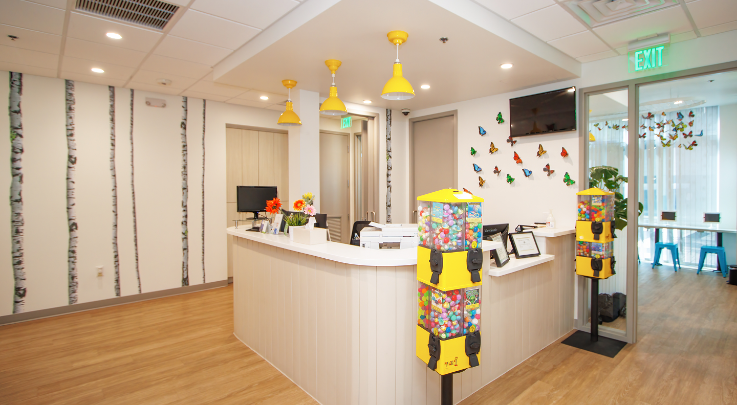 Tatum Pediatric Dentistry | A reception area with a white curved desk, yellow pendant lights, colorful gumball machines, and a wall decorated with vibrant butterfly decals.