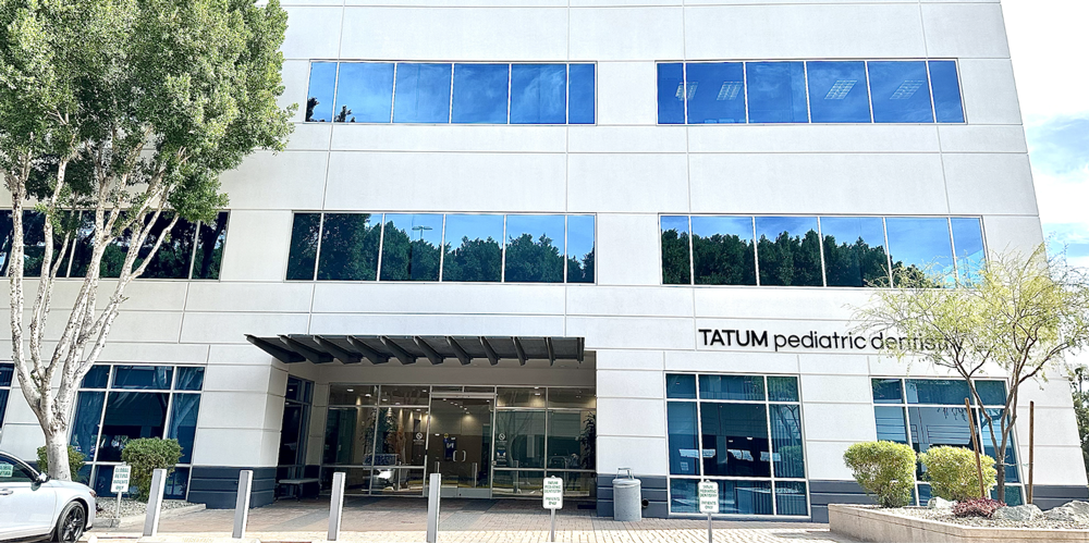 Tatum Pediatric Dentistry | Exterior view of the Tatum Pediatric Dentistry building, a modern white office structure with large glass windows reflecting trees and the sky.