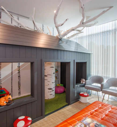 Tatum Pediatric Dentistry | Whimsical children's play area in a dental office, featuring a dark gray treehouse-like structure with white tree branch decor, small mushroom stools, and a cozy play space inside.