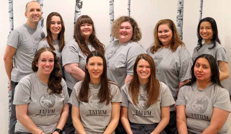 Tatum Pediatric Dentistry | Group photo of the Tatum Pediatric Dentistry team wearing matching gray t-shirts with the clinic's logo, posing against a birch tree-themed wall.