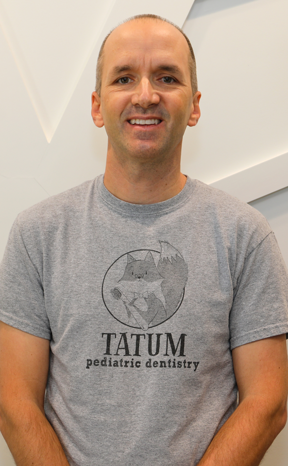 Tatum Pediatric Dentistry | Smiling man named Dr. Guymon wearing a gray 'Tatum Pediatric Dentistry' t-shirt, sitting against a light-colored geometric wall.