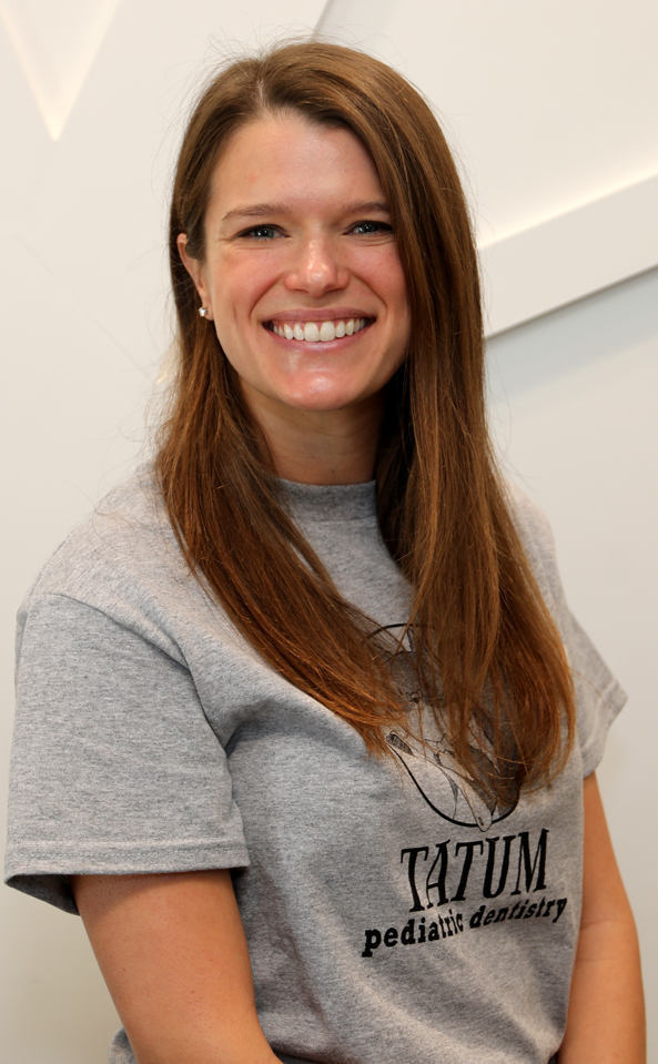 Tatum Pediatric Dentistry | Smiling woman named Dr. Carney with long brown hair wearing a gray 'Tatum Pediatric Dentistry' t-shirt, sitting against a light-colored geometric wall.